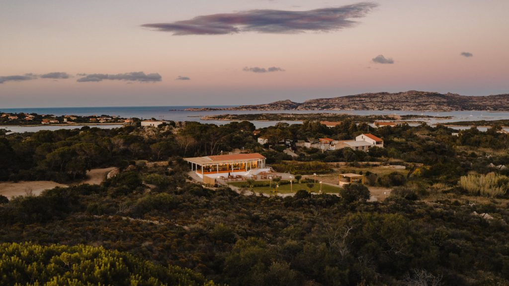 Veduta aerea del ristorante Kumale al tramonto sull'isola della Maddalena. L'edificio principale, con tetto in tegole rosse e illuminazione interna calda, è circondato da vegetazione mediterranea e si affaccia sul mare. Tavoli esterni sono visibili nel giardino. Lo sfondo mostra un paesaggio costiero frastagliato con altre abitazioni sparse e il cielo crepuscolare con nuvole rosate e grigie