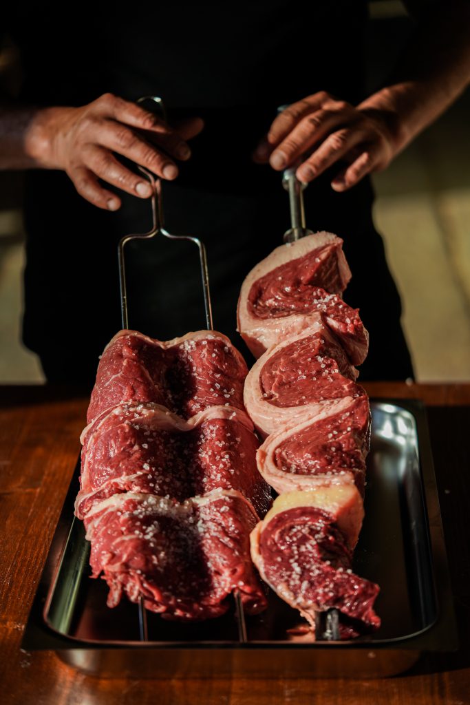 Due spiedi di carne cruda, presumibilmente picanha, infilzati e cosparsi di sale grosso, pronti per essere cotti. Uno spiedo presenta un pezzo di carne più grande e compatto, mentre l'altro mostra fette di carne avvolte a spirale con strati di grasso visibili. Le mani di un cuoco, che indossa una maglietta nera, tengono saldamente gli spiedi sopra una teglia di metallo. La superficie sottostante sembra essere un tavolo di legno