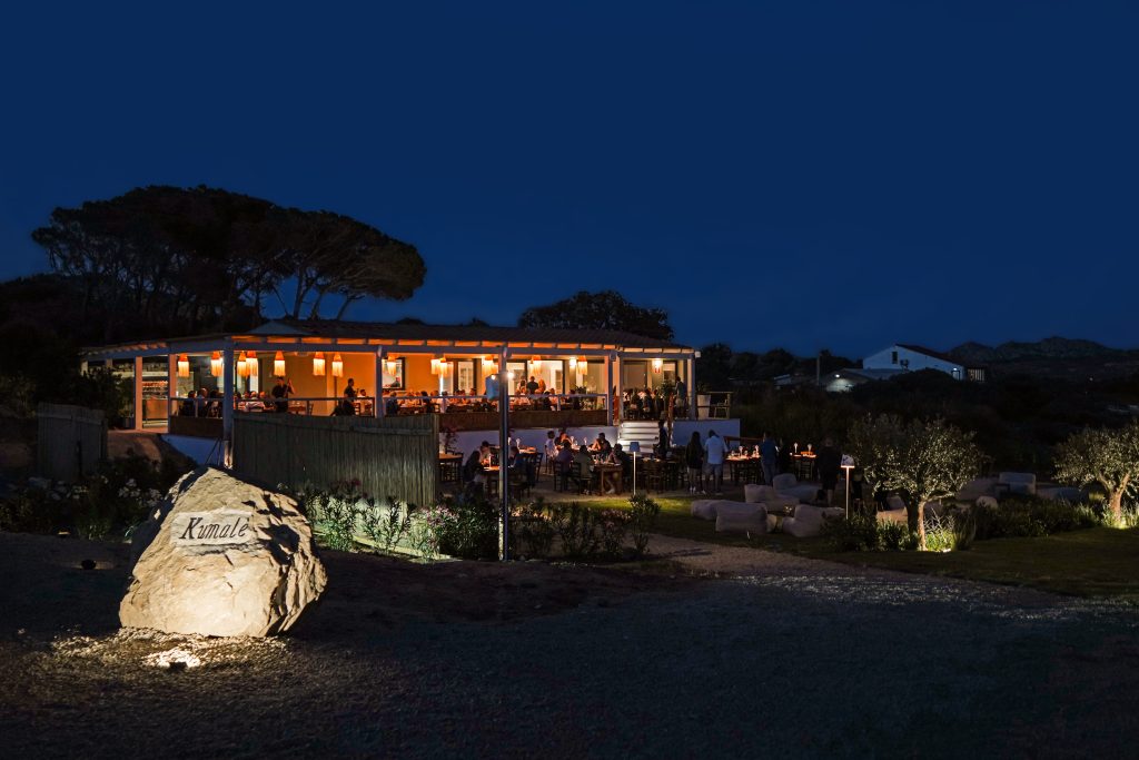 Veduta notturna del ristorante Kumale, una churrascaria brasiliana all'aperto sull'isola della Maddalena. L'edificio principale è illuminato con luci calde, mostrando clienti seduti ai tavoli all'interno. All'esterno, ulteriori tavoli sono disposti su una terrazza e nel giardino circostante, anch'essi illuminati. In primo piano a sinistra, una grande pietra con il nome "Kumale" inciso e illuminato da un faretto. L'atmosfera è accogliente e rilassata, con la vegetazione circostante visibile sullo sfondo sotto un cielo notturno blu scuro